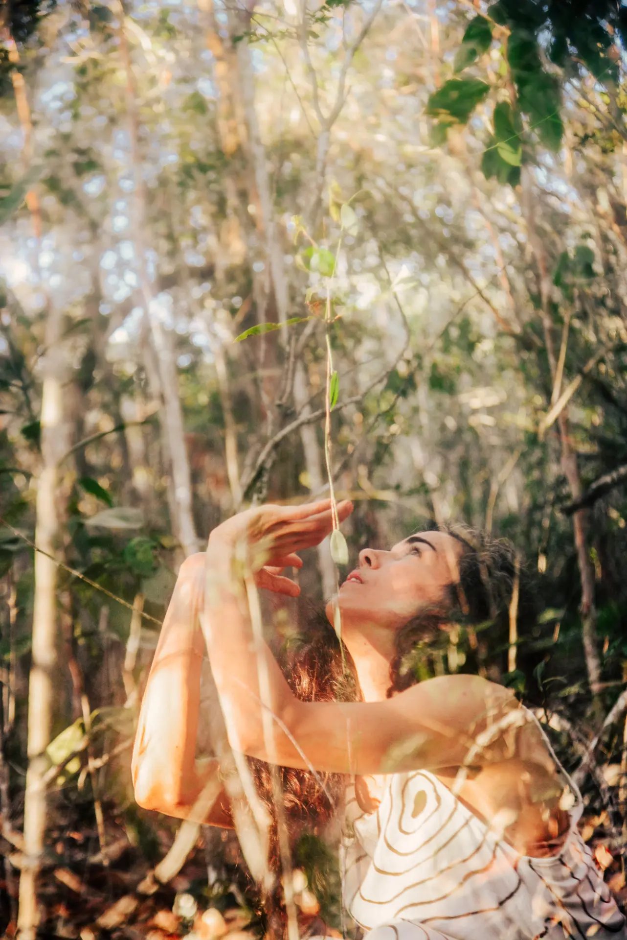 Juliana Lião em meio à mata, com os braços erguidos e o olhar voltado para cima, entre folhas e luz filtrada