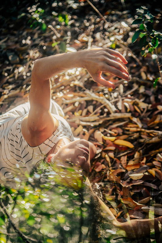 Juliana deitada na floresta entre folhas secas, com o braço estendido em um gesto de dança.